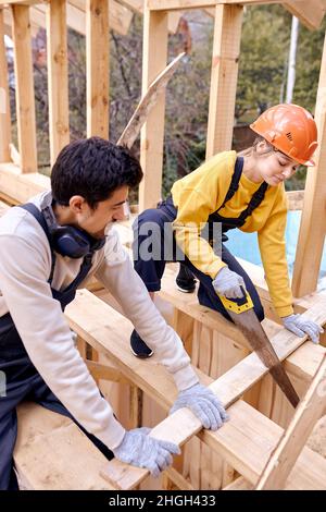Caucasian Contractors Carpenters Workers Using Wood Saw in Construction Zone. Wooden Skeleton Framing Building. Hardworking Cooperative Man And Woman Stock Photo