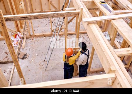 Top view on two caucasian engineers builders working together, building wooden cottage house. Young hardworking carpenters in special clothes work and Stock Photo