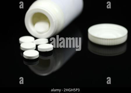 White medical pills and tablets pouring out of a medicine bottle on mirror black background. Blurred background. Macro view with copy space. Stock Photo