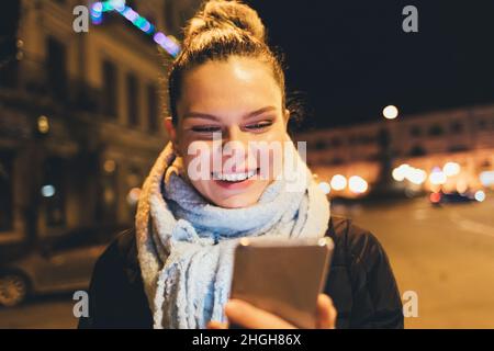 Portrait happy young woman using her smartphone walking on city street on winter evening Stock Photo