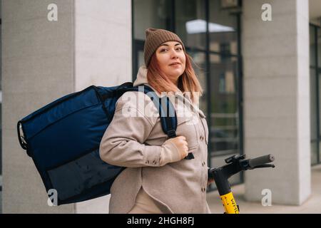 Side view of preofessional female courier with big thermal backpack standing with electric scooter in city street looking at camera. Stock Photo
