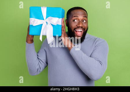 Photo of impressed african guy dressed grey sweater smiling guessing inside gift isolated green color background Stock Photo