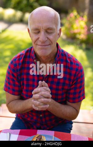 Caucasian senior man with eyes closed and hands clasped praying in garden Stock Photo
