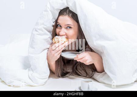 Cute woman is eating a cupcake while lying on the bed, under the covers. Breakfast in bed. Dessert. Calories. Stock Photo