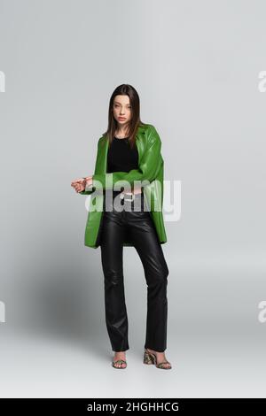 full length view of brunette woman in leather trousers and jacket standing on grey Stock Photo