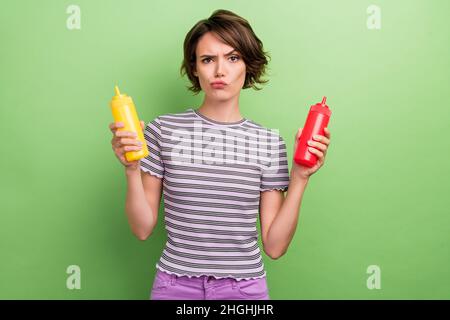 Photo of mad young brunette lady press ketchup mustard wear striped t-shirt isolated on green color background Stock Photo