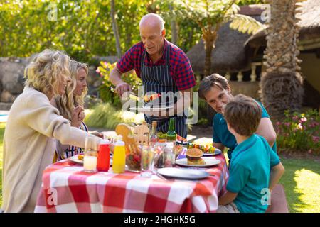 Caucasian senior man serving barbecue food to family sitting together at table in the garden Stock Photo