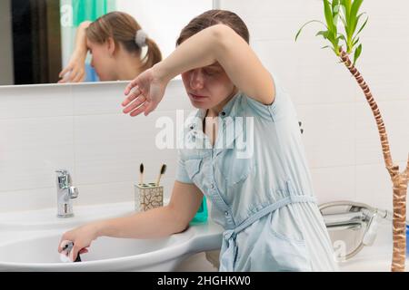 A young woman cleaner in a bright bathroom with a plunger to clear the blockage in her hands. On the wall is a mirror with a reflection. Green plant. Stock Photo