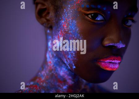 black female with neon make-up body art posing looking at camera, isolated on purple. beautiful female with dark skin with colourful paintings on face Stock Photo