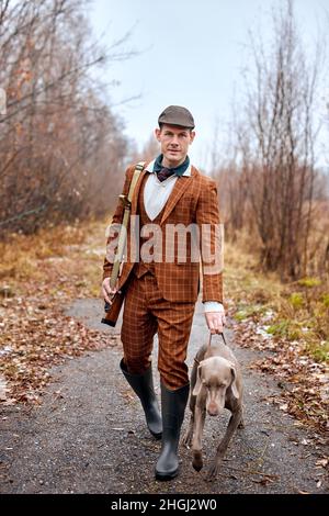 Young handsome male in suit hunter in forest, man holding gun and walking with dog. Hunter with shotgun gun on hunt. strong beautiful Dog waiting for Stock Photo
