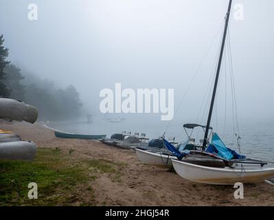 Sailboat on the beach of beautiful lake during a foggy morning. Other watercraft lined up along the sandy shore. Trees visible in background amidst th Stock Photo
