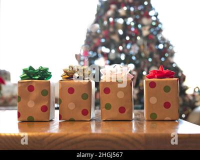 Four beautifully wrapped gift boxes on wood table with colorful bows, with decorated Christmas tree in background. Stock Photo
