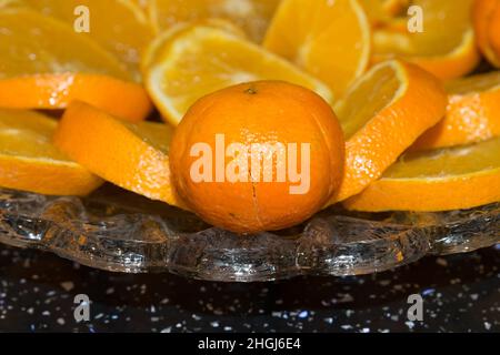 close up of bright orange sliced and whole oranges, Satsuma fruits on a pretty glass serving plate on a black kitchen table Stock Photo