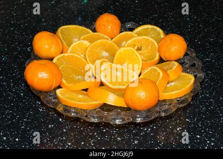 close up of bright orange sliced and whole oranges, Satsuma fruits on a pretty glass serving plate on a black kitchen table Stock Photo
