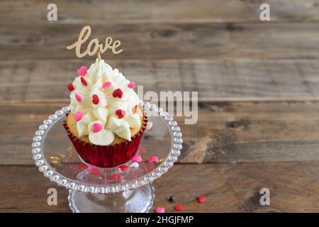 Cupcake on a display stand with cream and red topping decoration on a wooden background with a sign with the word LOVE. Valentine's Day. Copy space. Stock Photo