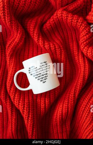 A white cup with a heart stands on a red knitted fabric. The concept of love and celebration of St. Valentine's day, February 14th Stock Photo