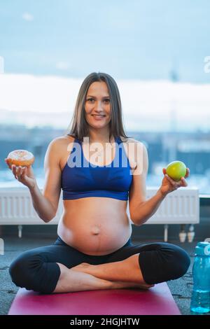 athletic pregnant woman in sportswear hold donut and apple sitting on yoga mat Stock Photo