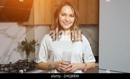 Woman drinks water from glass smiling in kitchen at home Stock Photo