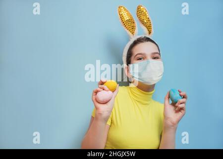 Teen holding decorated eggs. Schoolgirl with bunny ears Easter, beautiful girl in yellow suit on blue background. Copy space Stock Photo