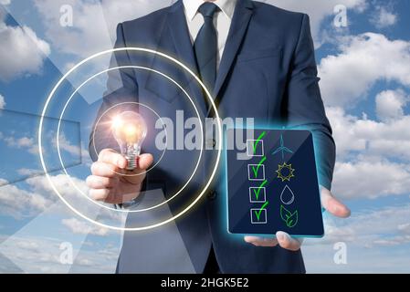 A man in a suit holds a lit light bulb while showing a panel with renewable energies. Caring for the environment Stock Photo