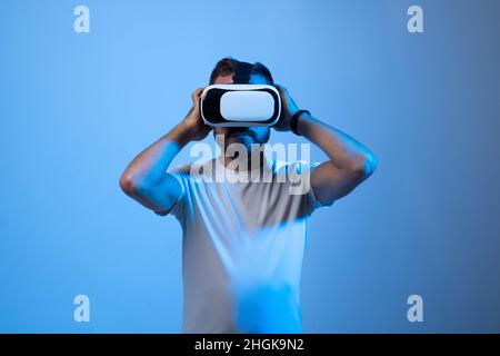 Young bearded guy in VR goggles touching air and excited with new feelings in dark neon lights. Stock Photo
