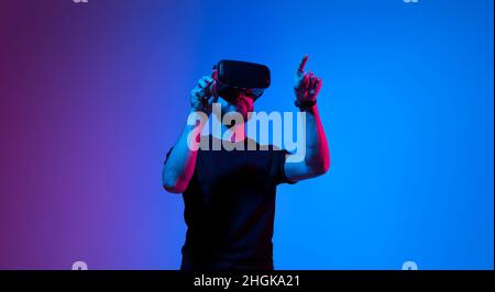 Young man uses virtual reality headset. Cheerful bearded man in t-shirt moving hands and using VR headset in neaon light background. Technology Stock Photo