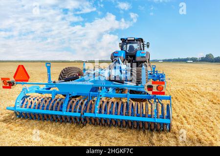 Trailed blue harrow as a trailed implement on a tractor against the background of yellow stubble and blue sky on a clear day. Stock Photo