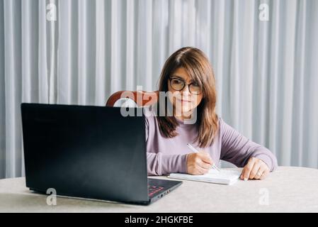 Hispanic woman with glasses looking at laptop while writing or taking notes on notebook with a pen Stock Photo