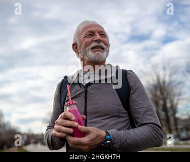 Senior bearded man in sportswear standing outdoor with backpack on his shoulders and holding fresh healthy juice in his hands Stock Photo