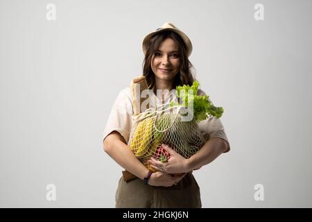 Portrait of happy smiling young woman in brown dress holding reusable string bag with groceries over orange background. Sustainability, eco living and Stock Photo