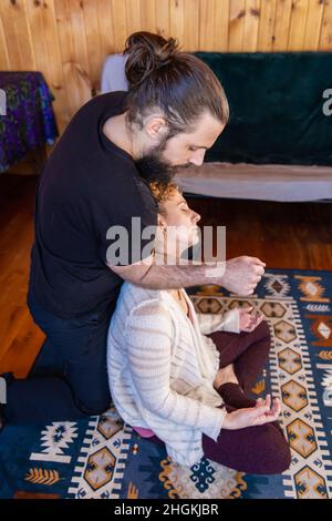 A male therapist with hair in bun is seen from the side, performing deep neck and shoulder massage to a relaxed woman sitting in the lotus position. Stock Photo