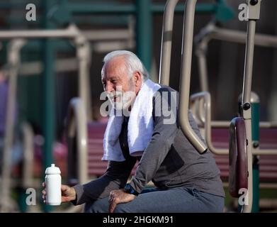 Senior men drinking water after using equipment in the outdoor public gym and wearing towel around his neck Stock Photo