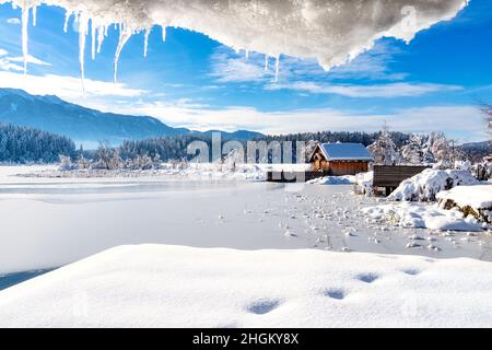 A panoramic view on the Faaker lake in Austrian Alps. The lake is surrounded by high mountains. There is a snow covered pier going into the frozen lak Stock Photo