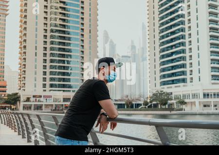 Young person in medical mask standing outdoor and looking forward to the urban background with city skyline and skyscrapers around the lake Stock Photo