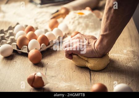 Hand of a man kneading dough by hand on a wooden surface with flour and eggs around. Stock Photo