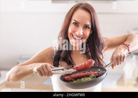 Smiling woman holds freshly pan-grilled sausage in pliers in her home kitchen. Stock Photo