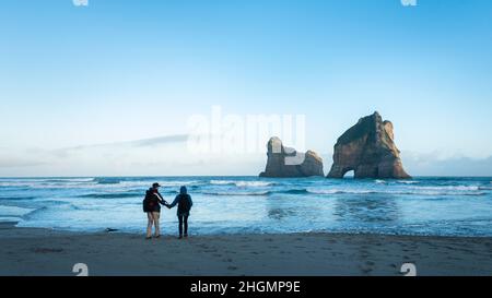 Couple standing on Wharariki Beach and watching Archway Island at Sunrise, South Island of New Zealand. Stock Photo