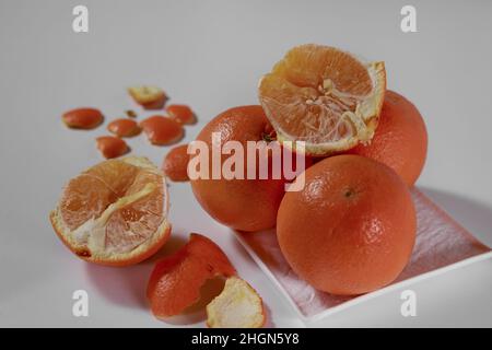 Four oranges lie on a white ceramic plate and one is broken in half with a peeled peel on a white background. Stock Photo