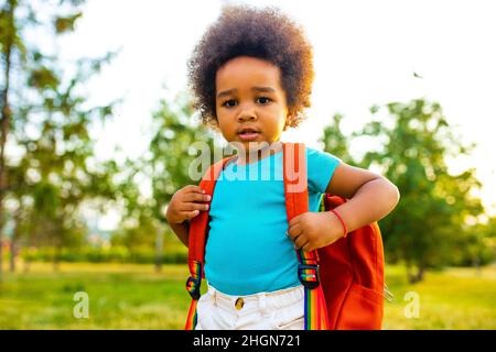 mixed race cute little girl with an orange backpack in blue cotton t-shirt looking at camera outdoors in park Stock Photo