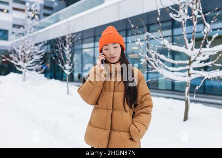 Young Asian woman talking on the phone walking on a snowy winter day, a student on campus Stock Photo