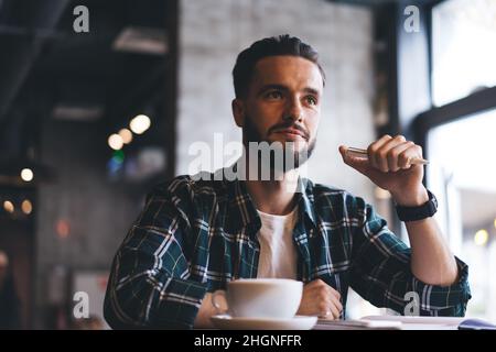 pensive hipster guy with caffeine cappuccino sitting at cafeteria table and feeling pondering Stock Photo
