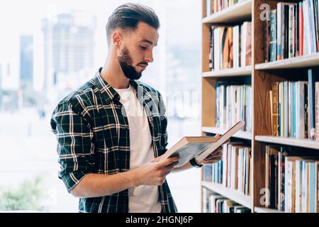 Young male librarian in casual clothing making order near bookshelves in college campus Stock Photo