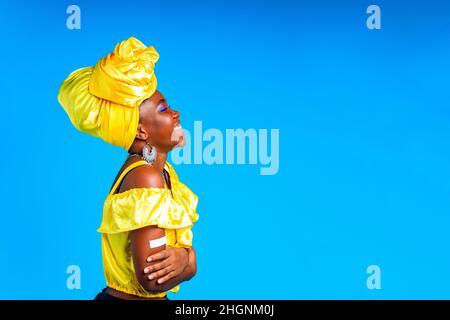 african woman in yellow silk dress blouse and turban over head showing her vaccinated arm in blue studio background Stock Photo