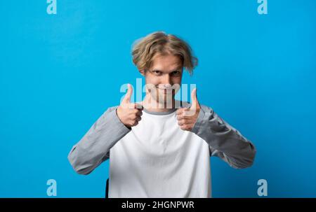 Young handsome blonde man in white t-shirt doing happy thumbs up gesture with both hand. Approving expression looking at the camera, showing success. Stock Photo
