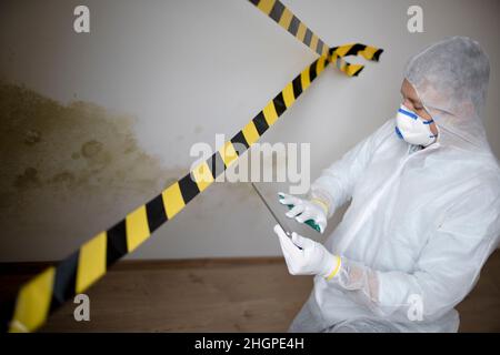 Man with white protective suit and mouth nose mask stands in front of mold on wall and works with tablet behind barrier Stock Photo