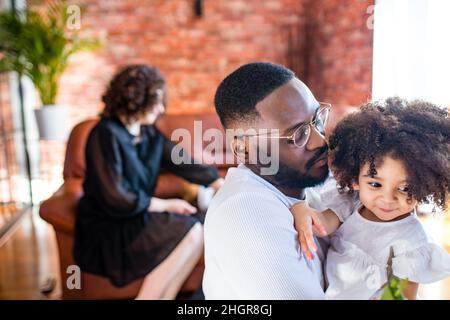 Portrait of upset man hugs his cute curly baby girl in living room Stock Photo