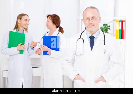 doctor posing in office, he is wearing a stethoscope, medical staff on the background. Quality medicine concept. Copy space Stock Photo