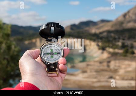 Hand of a hiker holding a compass to orient himself in a westerly direction. In the background is a river, blue sky and mountains out of focus.