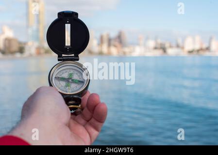 Hand holds a compass over the sea to orient in the city with an out-of-focus city skyline in the background. Stock Photo
