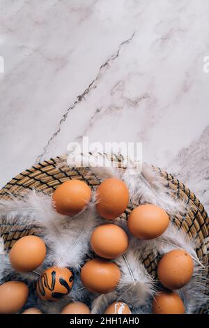 Top view composition of brown chicken eggs and white fluffy birds feathers on round wicker stand on marble table. Easter holiday decorations, flat lay Stock Photo